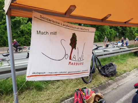 Das Solawi-Banner im Vordergrund, die Mobilitätsspur mit Fahrradfahrer*innen im Hintergrund.